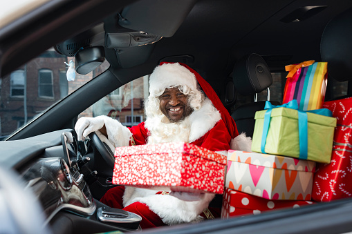 A medium shot of a senior man wearing a Santa Claus costume. He sits in his delivery van with a stack of Christmas presents on the seat beside him. He looks and smiles at the camera.