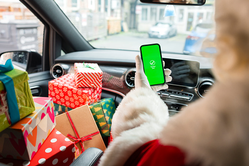 A medium, over-the-shoulder shot of a senior man wearing a Santa Claus costume. He sits in his delivery van with a stack of Christmas presents on the seat beside him. He is holding his smartphone showing a graphic which reads 