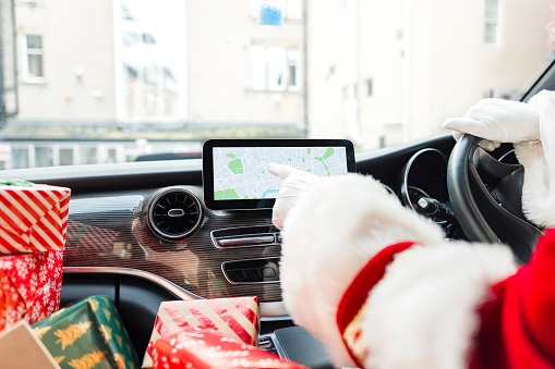 A medium, over-the-shoulder shot of a senior man wearing a Santa Claus costume. He sits in his delivery van with a stack of Christmas presents on the seat beside him. He is using the satellite navigation system in his van.