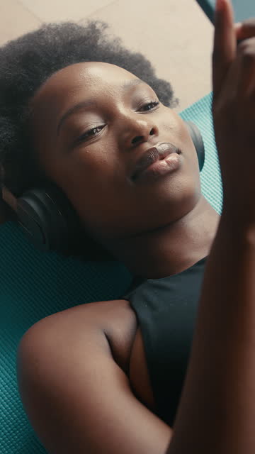 Relax, phone black woman with headphones on a gym floor for music, text or social media. Smartphone, app and African lady athlete at sports center after yoga, streaming or pilates fitness or workout