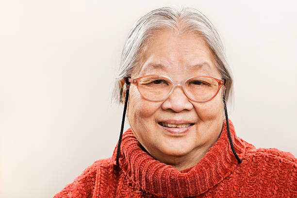 Asian Senior Woman in 80s, Cheerful, Happy Retired Chinese Grandmother An Asian senior woman, cheerful and smiling for the camera portrait. The happy, retired Chinese grandmother in her 80s has gray hair and wears reading glasses and a comfortable red sweater. Photographed against a plain light neutral background with copy space. grandma portrait stock pictures, royalty-free photos & images