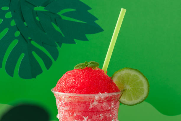 A strawberry and cherry red fruit flavored shaved ice or slush with lemon in a clear disposable cup on a green paper background decorated with green paper leaves.