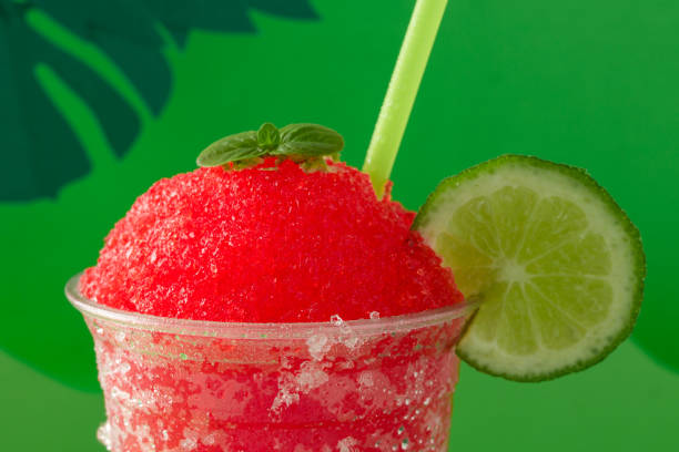 A strawberry and cherry red fruit flavored shaved ice or slush with lemon in a clear disposable cup on a green paper background decorated with green paper leaves.