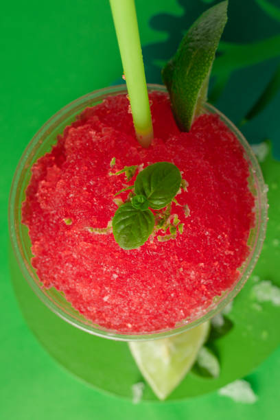 A strawberry and cherry red fruit flavored shaved ice or slush with lemon in a clear disposable cup on a green paper background decorated with green paper leaves.