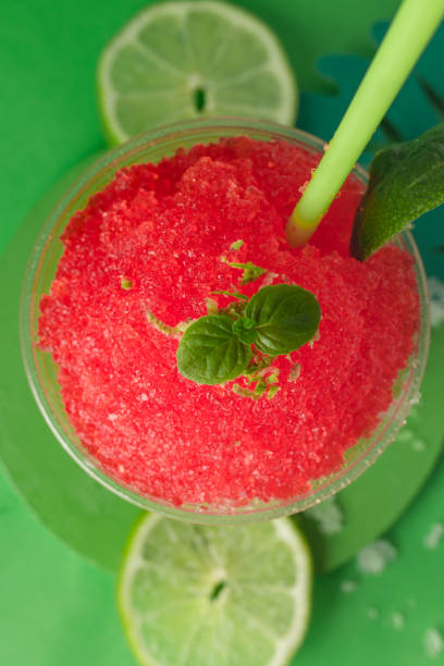 A strawberry and cherry red fruit flavored shaved ice or slush with lemon in a clear disposable cup on a green paper background decorated with green paper leaves.