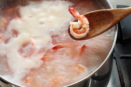 One boiled shrimp in a wooden spoon with a pot of boiling shrimp in the background.