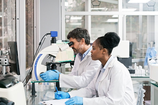 Young female biochemist sitting by mature male technician looking through microscope