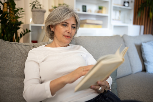 Mature woman reading a book while sitting on sofa in living room at home.