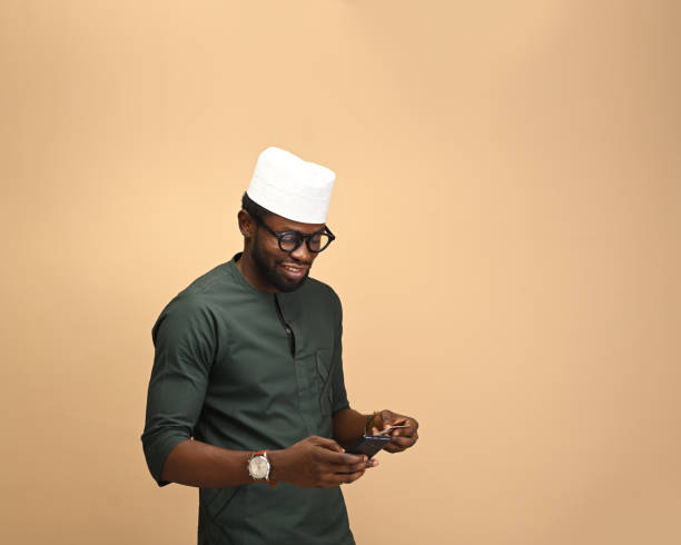 A traditional businessman from the African clan wearing brown attire and a cap to match, Indoors in a studio, standing by the Roadside, holding his smartphone and credit card. copy space. un hombre africano con su tarjeta de crédito pagando sus facturas y operaciones bancarias con su teléfono móvil y crédito. está usando su tarjeta de crédito para reservar un vuelo internacional. concepto de banca en línea y pago de facturas.