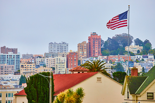 Image of American Flag in wind over residential rooftops with overcast sky and city behind it