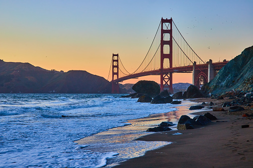 Image of Blue hour with glowing waves against sandy shore with distant Golden Gate Bridge