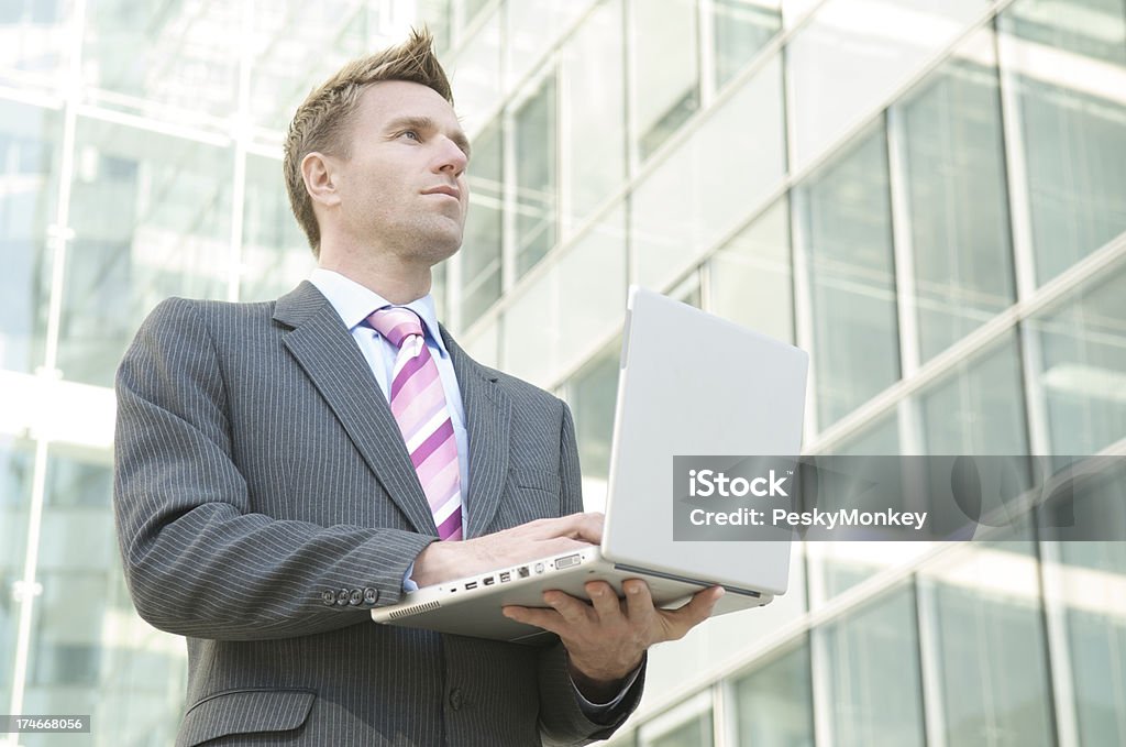 Businessman Stands in Corporate Courtyard Using Laptop Computer Optimistic businessman stands before a glass box background holding his laptop 30-39 Years Stock Photo