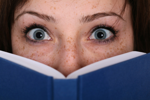 Young woman staring wide-eyed behind a scary book. She has freckles and deep blue eyes.