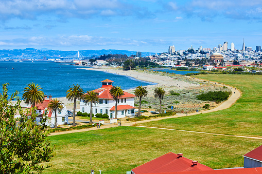 Image of Buildings on edge of beach with people on trail and distant San Francisco skyscrapers and bridge