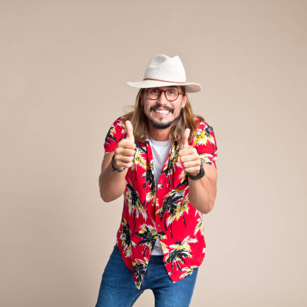 Portrait of excited man wearing red floral shirt and sun hat laughing at camera with thumbs up. Studio shot, beige background.