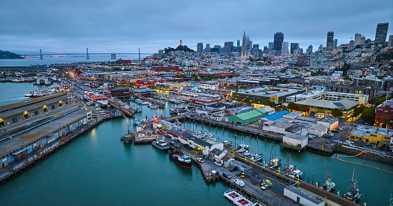 Image of Fishermans Wharf at dusk with night lights on over city and pier with Oakland Bay Bridge aerial