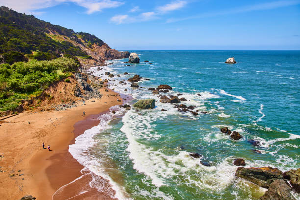 istock Green rocky coastline with sandy beach speckled with people watching crashing waves 1746716604