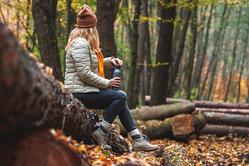 Refreshment during hiking in nature. Woman with thermos resting on log in autumn forest