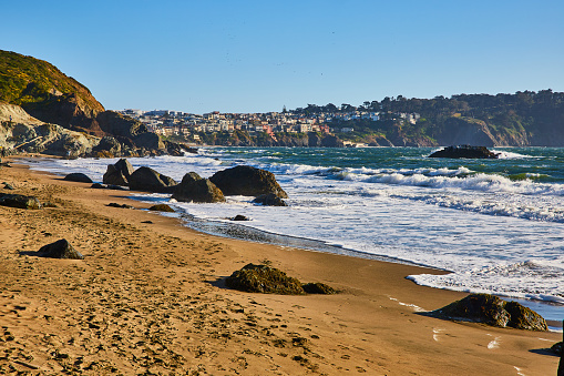 Image of Mossy boulders on beach with tons of footprints in sand and distant city above cliff and waves
