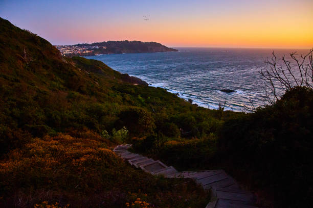 istock Steps going down hillside toward blue ocean with sunset sky and distant city above cliff 1746754671