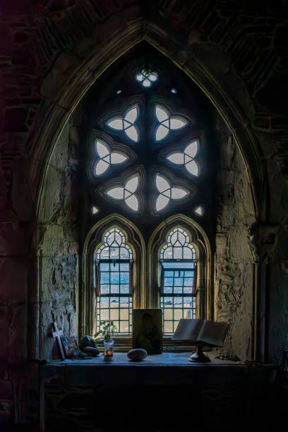 An ornate leaded glass window with a dimly lit informal alter set up on the sill. Looking through the window one can see the ocean and beach on a sunny day. Iona Abbey, Iona, Scotland