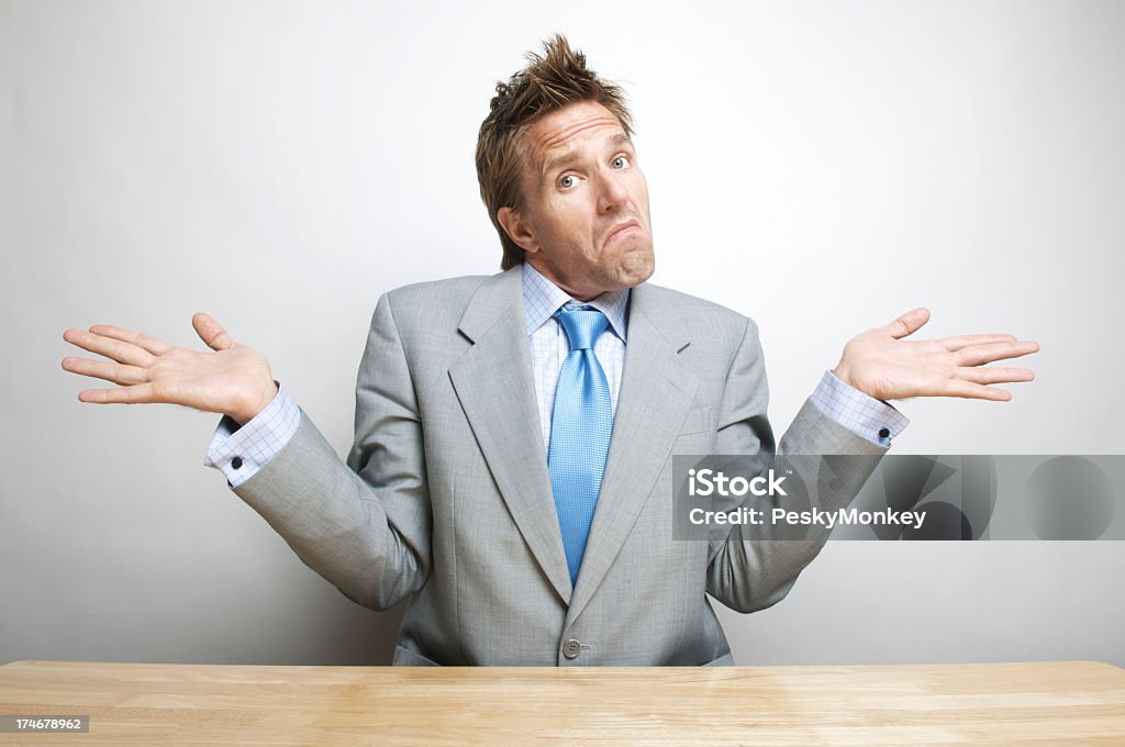 Doubtful Confused Office Worker Businessman Shrugs Shoulders at his Desk Dubious confused office worker businessman puts up his hands and shrugs sitting at his desk Shrugging Stock Photo