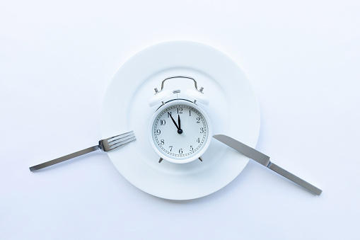 Directly above view of white plate with alarm clock and   fork and knife  on white background. Representing diet, Intermittent fasting, time-restricted eating and healthy food concept.