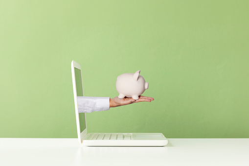 Hand holding white piggy bank  is reaching out of white laptop screen on white table in front of green wall. Representing online personal finance consultancy
