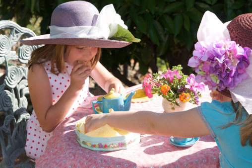 Two little girls wearing fancy hats are having a tea party outdoors.  They are busy eating cookies.Find more tea party photos here