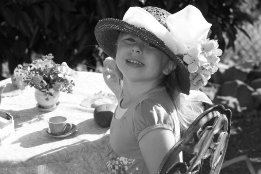 A cute little girl is looking at the camera smiling while wearing a big fancy hat at an outdoor tea party.Find more tea party photos here