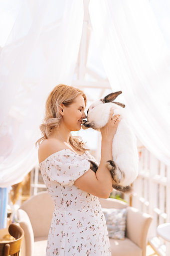 Vertical shot of happy blonde young woman in summer dress holding hugging cute fluffy white bunny standing inside of gazebo house on sunny day. Concept of excursion to eco-farm, life in village.