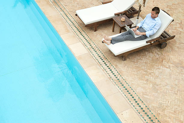 Businessman sits poolside in deck chair typing on his laptop