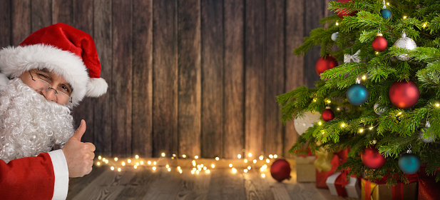 Winking Santa shows thumbs up and presents your text or content on a dark wood background copyspace between himself, a Christmas tree and glowing lights