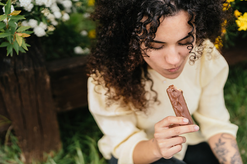 latin woman with curly hair enjoys chocolate bar in park