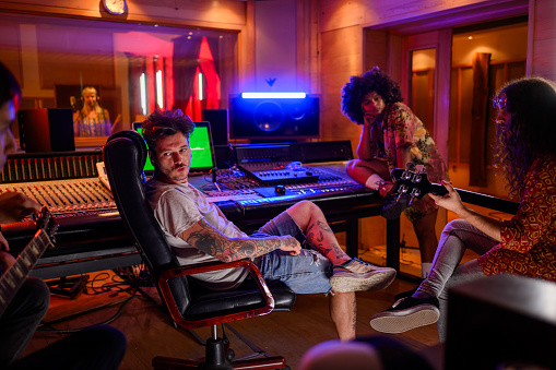 Photo of band members and a producer chilling out in a music studio while the vocalist is recording their track. They are relaxed and enjoying the process. Bass player is jamming and the guitarist is tuning the guitar.