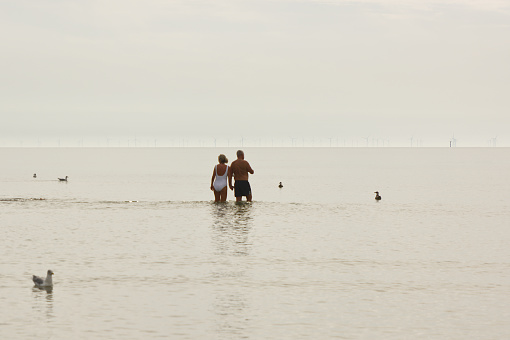 Man and woman walking in very calm sea with white sky and mist. Littlehampton, West Sussex, England