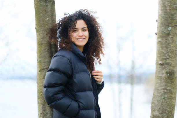 istock young curly haired woman leaning on tree and wearing coat smiles looking at camera 1746864972