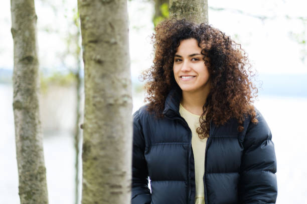 istock young curly haired woman leaning on tree and wearing coat smiles looking at camera 1746866194