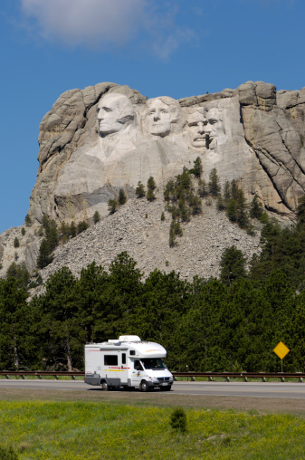 RV on road in foreground of Mt. Rushmore with blue sky and clouds.