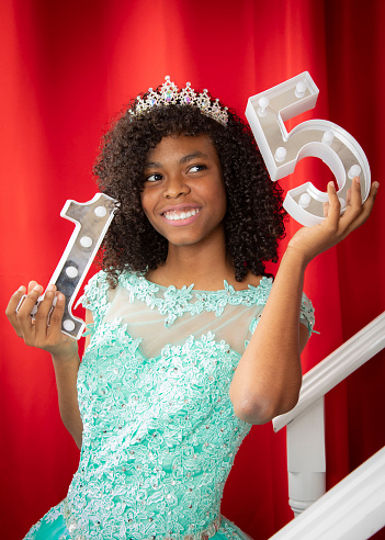 Hispanic girl celebrating her sweet 15th in a pick background wearing a blue dress looking as a princess with a tiara and holding the number 15 in her hands