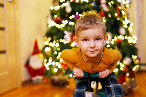 Happy little child boy sitting on push bicycle on a Christmas tree background. Christmas Eve