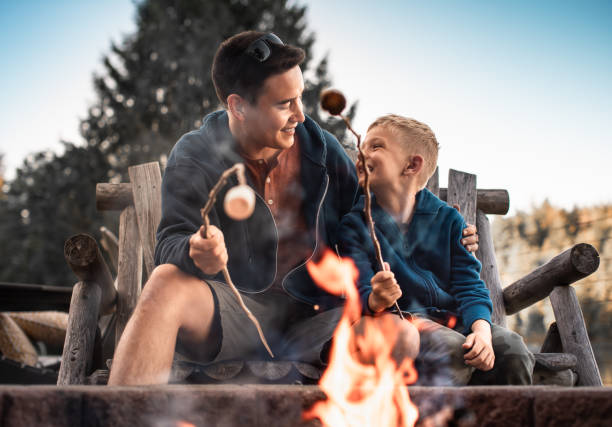Happy father and son enjoying time together camping, roasting marshmallows talking smiling bonding.