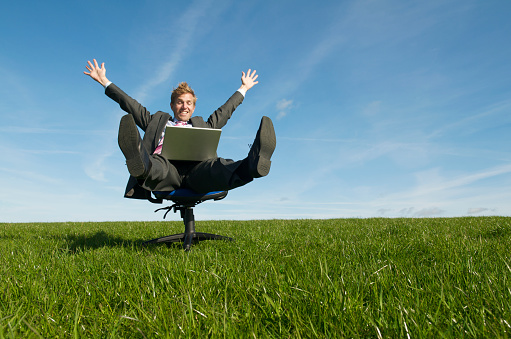 Excited happy businessman celebrating success spinning with his laptop outdoors on an office chair on a green meadow