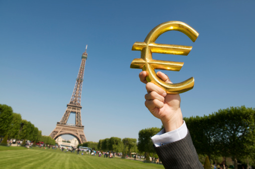 Hand of a businessman holds up a golden euro symbol in the blue sky next to the Eiffel Tower
