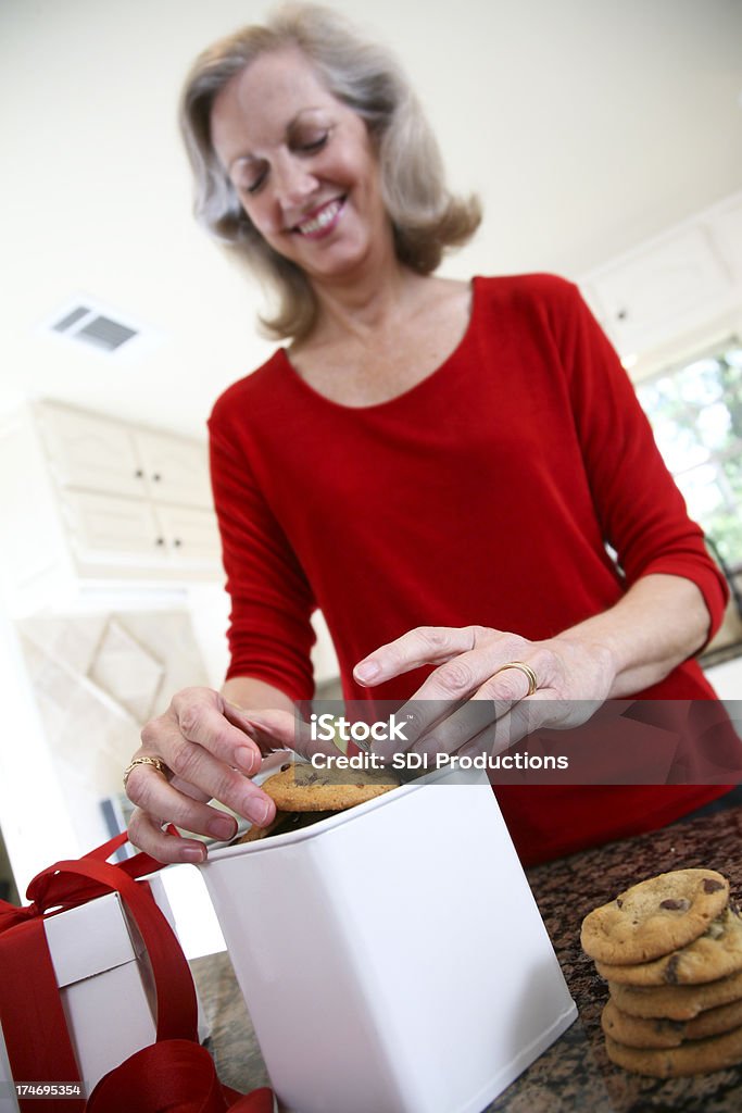 Smiling Woman Placing Chocolate Chip Cookies in Gift Box Smiling Woman Placing Chocolate Chip Cookies in Gift Box. Focus is on the Cookies.See more from this series: 50-59 Years Stock Photo
