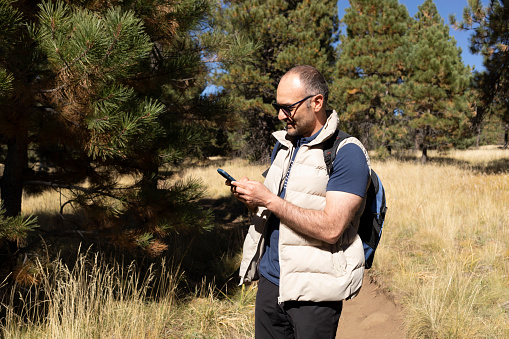Smiling 40 yo Man in Sunglasses Holding Smartphone, Gadget. Fir Trees, Forest On Background. Nature in Fall Leaves Season. Horizontal. Active Lifestyle. Male Trekking or Hiking. Copy Space