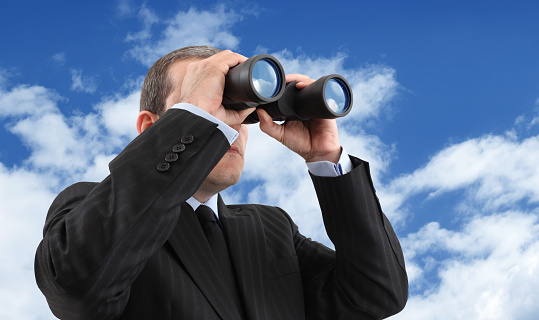 Businessman looking through a binoculars with a reflection of a blue and cloudy sky.
