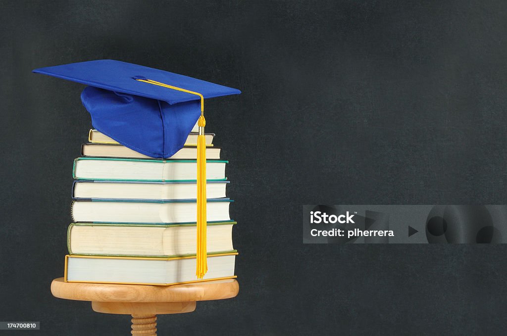 Blue and yellow graduation cap on book stack near chalkboard Blue graduation cap on a stack of books in front of a chalkboard. Back to School Stock Photo