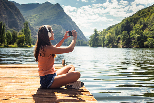 Shot of happy woman listening to music with headphones while taking photos with smartphone to the lake