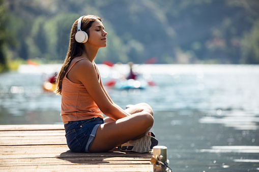 Shot of sporty woman listening to music with headphones while relaxing sitting by a lake